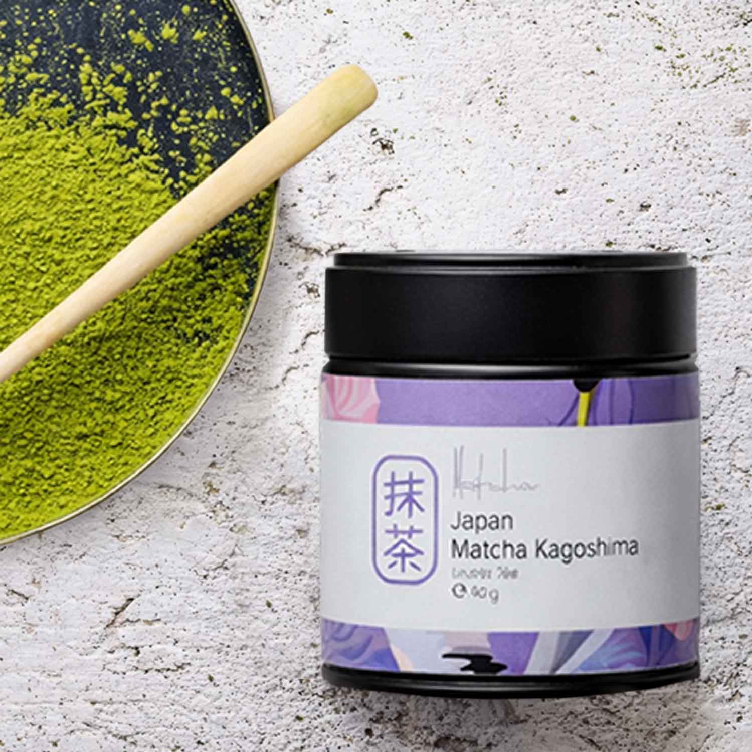 Matcha Kagoshima - Bio-Matcha für einen harmonischen Einstieg Schwarze Dose japanischen Matcha-Kagoshima-Tees neben einem Bambusbesen und einem kleinen Haufen leuchtend grünem Matcha-Pulver auf einem Tablett.